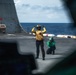 The image shows two naval personnel on the flight deck of a warship at sea. One person wearing a yellow vest and helmet is giving hand signals with both arms raised, while a second person in a green jersey and helmet crouches nearby, appearing to observe or photograph. The scene is viewed through what appears to be a cockpit window or aircraft frame in the foreground.