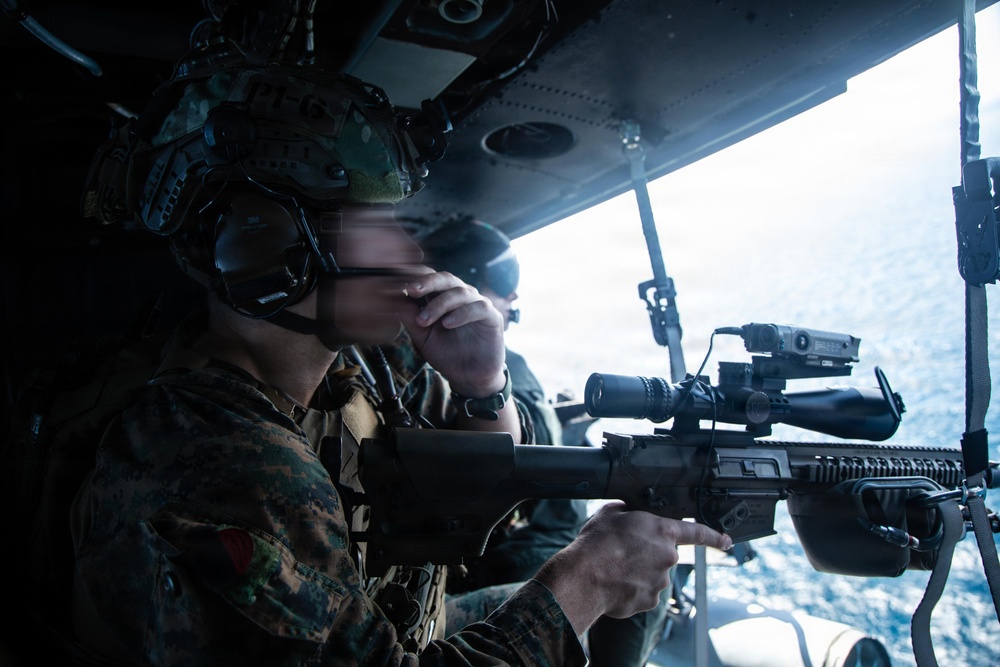 A military service member in camouflage uniform and tactical helmet is positioned inside what appears to be a helicopter or aircraft, aiming a rifle equipped with a scope and accessory rail out of an open door or window. The ocean is visible in the background, suggesting an airborne maritime operation. At least one additional personnel is visible in the background.