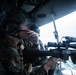 A military service member in camouflage uniform and tactical helmet is positioned inside what appears to be a helicopter or aircraft, aiming a rifle equipped with a scope and accessory rail out of an open door or window. The ocean is visible in the background, suggesting an airborne maritime operation. At least one additional personnel is visible in the background.