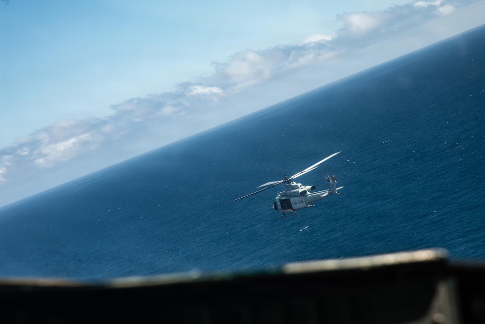 A military or coast guard helicopter is photographed in flight over open ocean, captured from an elevated vantage point such as a ship's deck or another aircraft. The image is taken at a tilted angle, giving the horizon a diagonal orientation. The helicopter appears to be white/light gray and is flying at low-to-medium altitude above deep blue water.