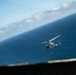 A military or coast guard helicopter is photographed in flight over open ocean, captured from an elevated vantage point such as a ship's deck or another aircraft. The image is taken at a tilted angle, giving the horizon a diagonal orientation. The helicopter appears to be white/light gray and is flying at low-to-medium altitude above deep blue water.
