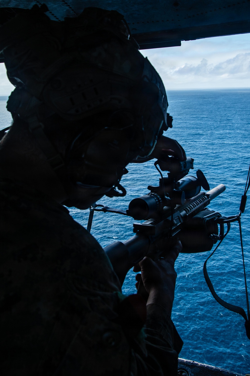 A silhouetted military personnel is positioned at the open door of an aircraft over open ocean, aiming a rifle equipped with an optical scope and rail-mounted accessories. The image is dramatically backlit, creating a strong contrast between the dark foreground figure and the bright blue ocean and sky in the background. The setting suggests an airborne maritime operation or training exercise.