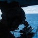 A silhouetted military personnel is positioned at the open door of an aircraft over open ocean, aiming a rifle equipped with an optical scope and rail-mounted accessories. The image is dramatically backlit, creating a strong contrast between the dark foreground figure and the bright blue ocean and sky in the background. The setting suggests an airborne maritime operation or training exercise.