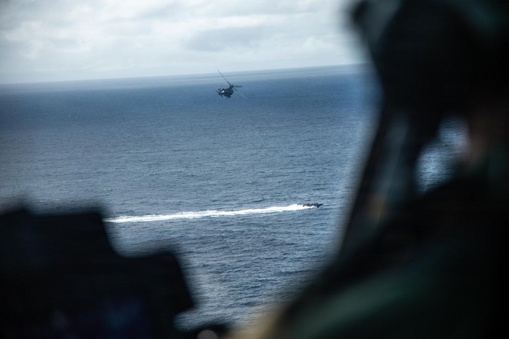 The image is taken from inside a military helicopter, showing a crew member in the foreground observing operations over open ocean. In the mid-distance, another helicopter hovers above the water while a fast-moving tactical boat leaves a white wake on the surface below. The scene appears to depict a coordinated maritime military exercise or operation.