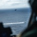 The image is taken from inside a military helicopter, showing a crew member in the foreground observing operations over open ocean. In the mid-distance, another helicopter hovers above the water while a fast-moving tactical boat leaves a white wake on the surface below. The scene appears to depict a coordinated maritime military exercise or operation.