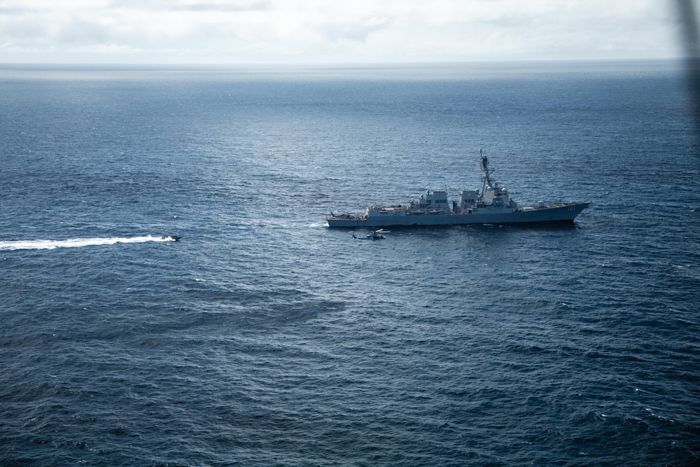 An aerial photograph taken from another aircraft showing a U.S. Navy Arleigh Burke-class guided-missile destroyer underway in open ocean. A military helicopter is visible hovering or flying low near the ship's bow, and a small fast-moving vessel is creating a wake to the port side of the destroyer. The scene appears to depict a naval exercise or operational activity at sea.