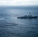 An aerial photograph taken from another aircraft showing a U.S. Navy Arleigh Burke-class guided-missile destroyer underway in open ocean. A military helicopter is visible hovering or flying low near the ship's bow, and a small fast-moving vessel is creating a wake to the port side of the destroyer. The scene appears to depict a naval exercise or operational activity at sea.