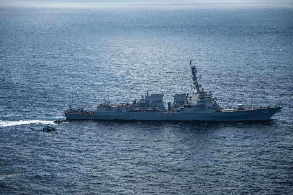 An aerial photograph shows a U.S. Navy Arleigh Burke-class guided-missile destroyer bearing hull number 101 underway at sea. A military helicopter is flying low alongside the vessel, and a small rigid-hull inflatable boat (RHIB) appears to be operating near the ship's port side. The image was taken from an elevated aerial vantage point over open ocean under overcast lighting conditions.