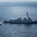 An aerial photograph shows a U.S. Navy Arleigh Burke-class guided-missile destroyer bearing hull number 101 underway at sea. A military helicopter is flying low alongside the vessel, and a small rigid-hull inflatable boat (RHIB) appears to be operating near the ship's port side. The image was taken from an elevated aerial vantage point over open ocean under overcast lighting conditions.