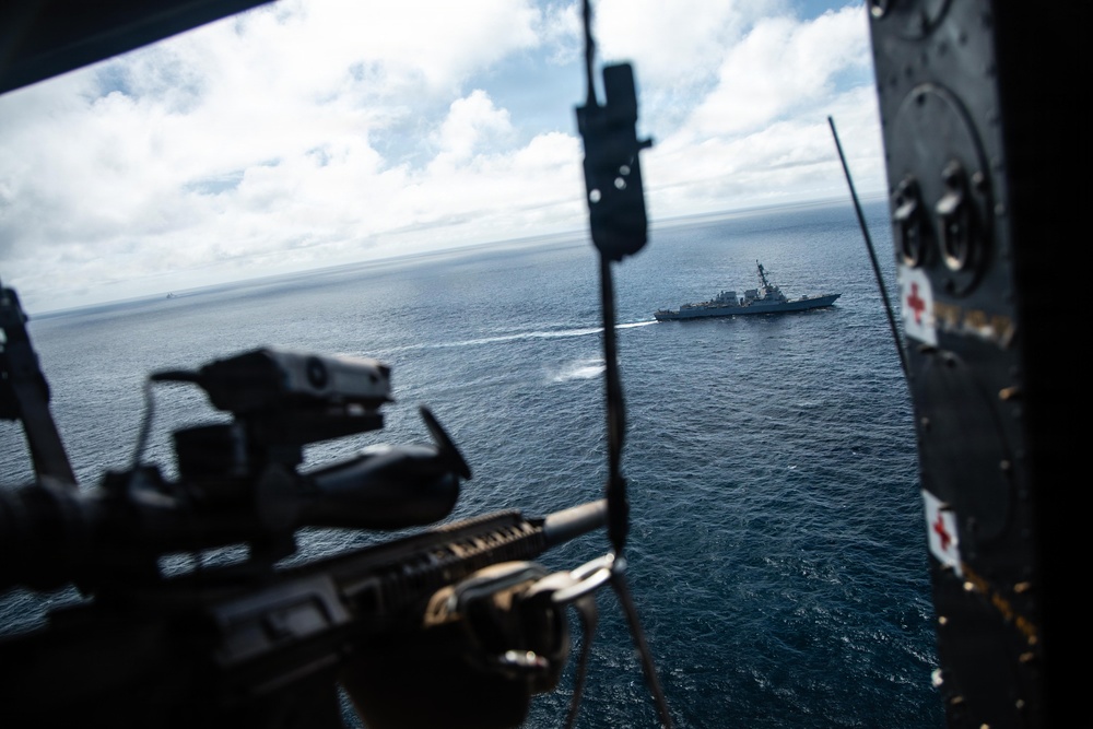 The image is taken from the open door or ramp of a military helicopter, looking out over open ocean toward a naval warship in the distance. A mounted machine gun is visible in the foreground, suggesting this is a combat or patrol helicopter conducting maritime operations. A red cross medical symbol is visible on the interior wall of the aircraft.