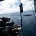 The image is taken from the open door or ramp of a military helicopter, looking out over open ocean toward a naval warship in the distance. A mounted machine gun is visible in the foreground, suggesting this is a combat or patrol helicopter conducting maritime operations. A red cross medical symbol is visible on the interior wall of the aircraft.