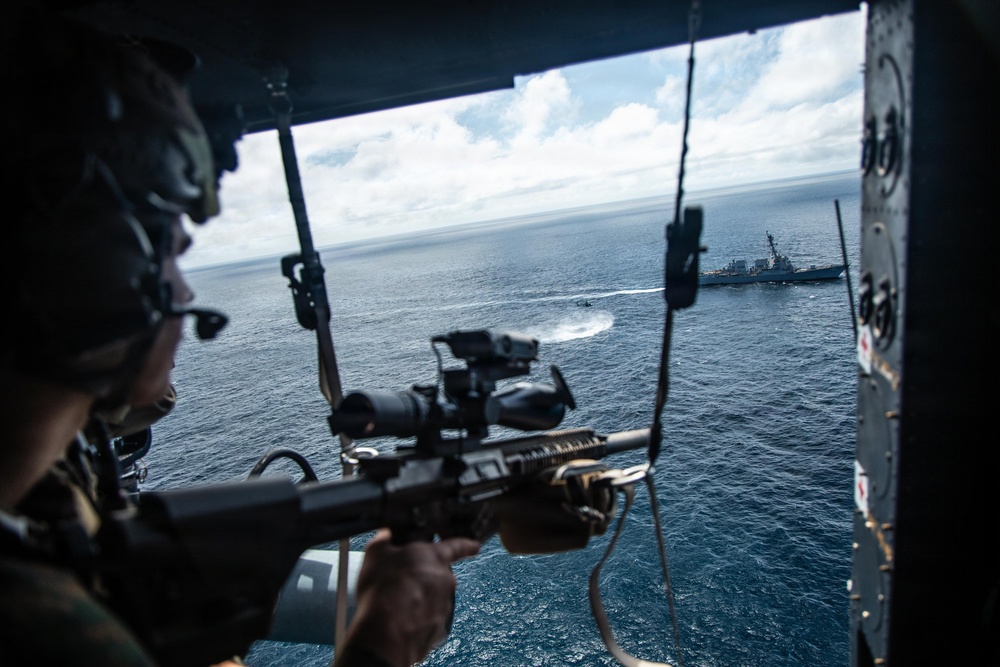 A military personnel is positioned inside a helicopter, aiming a scoped rifle toward the open ocean. In the background, a naval warship (appearing to be a destroyer) is visible on the water, along with what appears to be a smaller fast-moving vessel leaving a white wake. The scene appears to depict a maritime military exercise or operation.