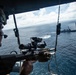 A military personnel is positioned inside a helicopter, aiming a scoped rifle toward the open ocean. In the background, a naval warship (appearing to be a destroyer) is visible on the water, along with what appears to be a smaller fast-moving vessel leaving a white wake. The scene appears to depict a maritime military exercise or operation.