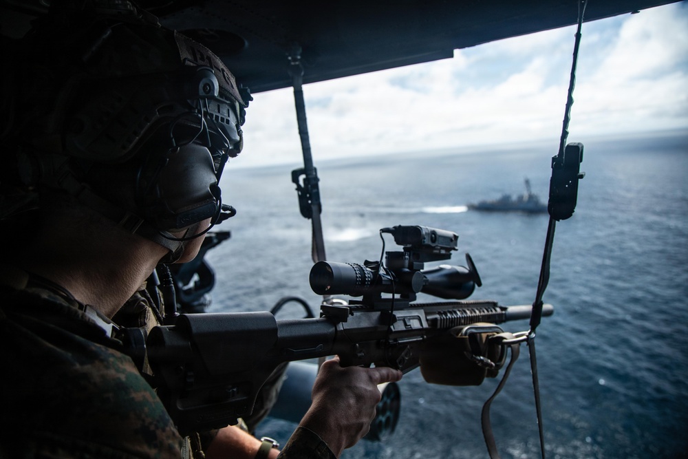 A military service member in combat gear is aiming a scoped rifle from what appears to be a helicopter doorway over open ocean. A naval vessel is visible in the background on the water below. The image appears to depict an aerial maritime security or training operation.