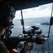 A military service member in combat gear is aiming a scoped rifle from what appears to be a helicopter doorway over open ocean. A naval vessel is visible in the background on the water below. The image appears to depict an aerial maritime security or training operation.