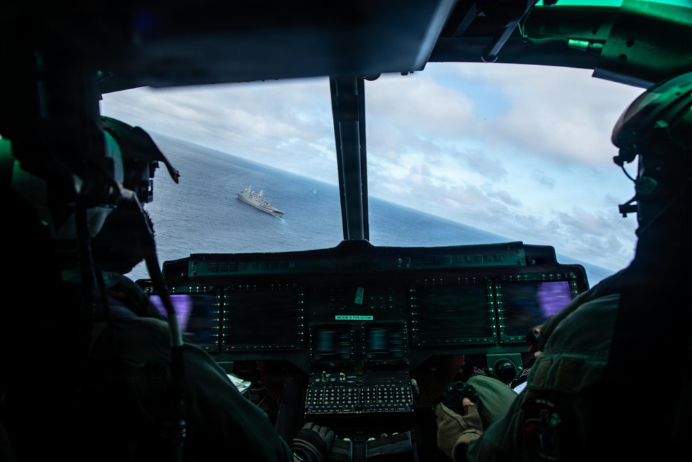 A view from inside a military helicopter or aircraft cockpit looking out over open ocean toward a naval warship below. Two crew members in flight helmets and gear are visible in the foreground operating the aircraft. The cockpit features modern glass displays with green-lit instrument panels.