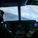 A view from inside a military helicopter or aircraft cockpit looking out over open ocean toward a naval warship below. Two crew members in flight helmets and gear are visible in the foreground operating the aircraft. The cockpit features modern glass displays with green-lit instrument panels.