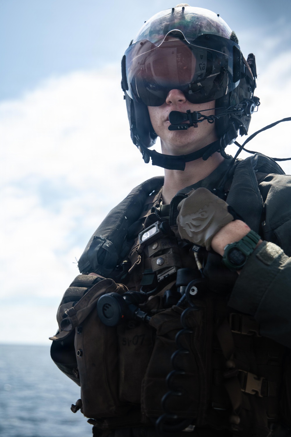 A military aviator or aircrew member is photographed in full flight gear against an open ocean and sky background. The subject is wearing a flight helmet with visor, communications equipment, tactical vest, and gloves. The image appears to be taken aboard a naval vessel given the visible ocean horizon.