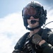 A military aviator or aircrew member is photographed in full flight gear against an open ocean and sky background. The subject is wearing a flight helmet with visor, communications equipment, tactical vest, and gloves. The image appears to be taken aboard a naval vessel given the visible ocean horizon.