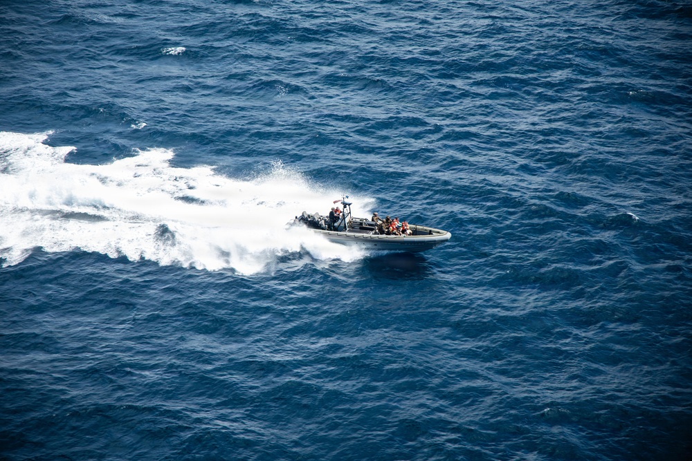 An aerial photograph shows a rigid-hull inflatable boat (RHIB) traveling at high speed across open deep-blue ocean water. The boat is leaving a prominent white wake and foam trail behind it. Several personnel are visible aboard the vessel, suggesting a military or law enforcement operation.