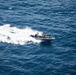 An aerial photograph shows a rigid-hull inflatable boat (RHIB) traveling at high speed across open deep-blue ocean water. The boat is leaving a prominent white wake and foam trail behind it. Several personnel are visible aboard the vessel, suggesting a military or law enforcement operation.