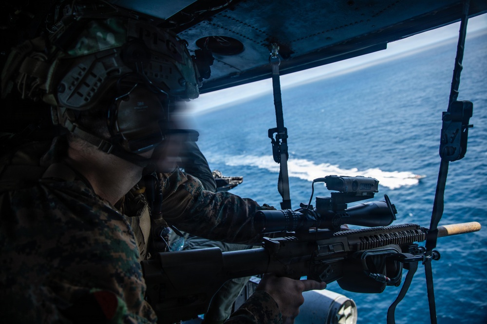 A military service member in MARPAT camouflage uniform and combat helmet is positioned at the open door of an aircraft, aiming a rifle equipped with optics and accessories toward the ocean below. The photograph is taken from inside the aircraft, showing a body of water with a visible wake trail from a vessel in the background. The image captures what appears to be an aerial gunnery or overwatch exercise conducted from a helicopter.