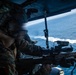 A military service member in MARPAT camouflage uniform and combat helmet is positioned at the open door of an aircraft, aiming a rifle equipped with optics and accessories toward the ocean below. The photograph is taken from inside the aircraft, showing a body of water with a visible wake trail from a vessel in the background. The image captures what appears to be an aerial gunnery or overwatch exercise conducted from a helicopter.