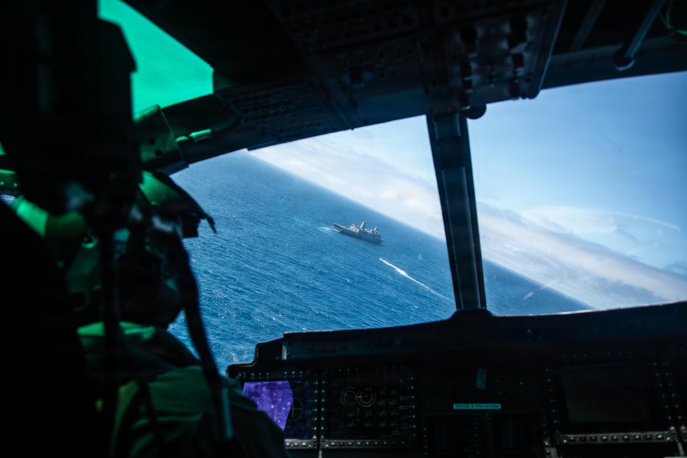 The image is taken from inside a military aircraft cockpit, looking through the windshield at a naval vessel sailing on open blue ocean below. A crew member in flight gear is partially visible on the left side of the frame. The cockpit instrumentation and structural framing are visible in the foreground, with the ship appearing small in the distance against a blue sea and partly cloudy horizon.