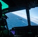 The image is taken from inside a military aircraft cockpit, looking through the windshield at a naval vessel sailing on open blue ocean below. A crew member in flight gear is partially visible on the left side of the frame. The cockpit instrumentation and structural framing are visible in the foreground, with the ship appearing small in the distance against a blue sea and partly cloudy horizon.