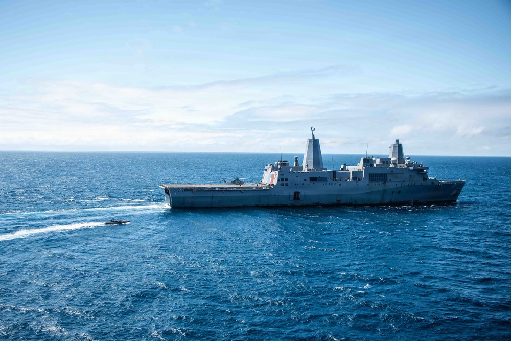 A large US Navy amphibious transport dock ship is underway on open ocean under partly cloudy skies. A smaller rigid-hull inflatable boat (RHIB) or patrol craft is visible operating nearby, leaving a visible wake. The ship displays a hull number visible on its bow and features a flat flight deck with superstructure amidships.