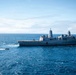 A large US Navy amphibious transport dock ship is underway on open ocean under partly cloudy skies. A smaller rigid-hull inflatable boat (RHIB) or patrol craft is visible operating nearby, leaving a visible wake. The ship displays a hull number visible on its bow and features a flat flight deck with superstructure amidships.