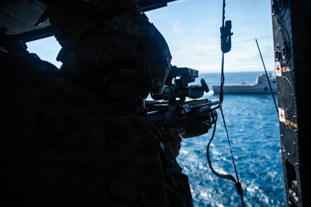 A military personnel in combat gear and helmet is aiming a rifle equipped with an optical scope from inside what appears to be a military helicopter or aircraft. The scene is set over open ocean water, with a large naval vessel visible in the background. The interior of the aircraft shows safety equipment markings on the right side panel.
