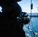 A military personnel in combat gear and helmet is aiming a rifle equipped with an optical scope from inside what appears to be a military helicopter or aircraft. The scene is set over open ocean water, with a large naval vessel visible in the background. The interior of the aircraft shows safety equipment markings on the right side panel.