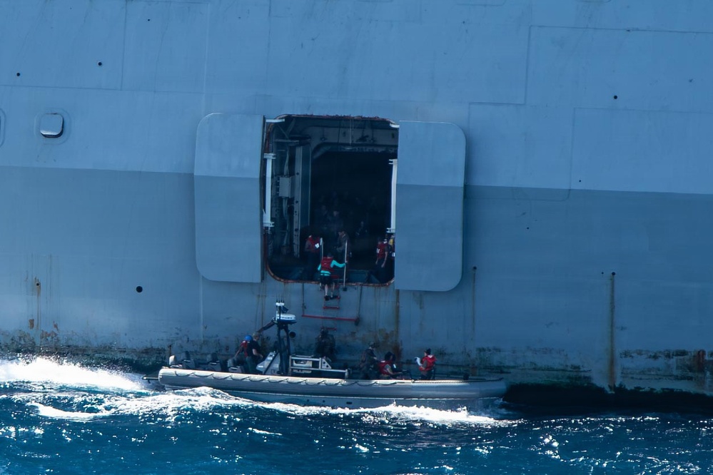 A rigid inflatable boat (RIB) is positioned alongside a large gray naval or military vessel at an open hull door/hatch at the waterline. Several personnel wearing life vests are visible both on the RIB and in the open hatch of the larger vessel, suggesting a transfer or boarding operation in progress. The sea conditions appear moderately choppy with visible white water around the smaller craft.