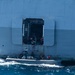 A rigid inflatable boat (RIB) is positioned alongside a large gray naval or military vessel at an open hull door/hatch at the waterline. Several personnel wearing life vests are visible both on the RIB and in the open hatch of the larger vessel, suggesting a transfer or boarding operation in progress. The sea conditions appear moderately choppy with visible white water around the smaller craft.