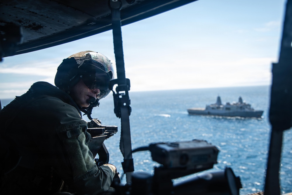 A military aircrew member wearing a flight helmet and tactical gear is positioned at an open door or window of an aircraft, appearing to operate or observe through a weapon or optical device. In the background, a naval vessel — consistent with a U.S. Navy amphibious transport dock ship — is visible on the open ocean. The image appears to document an aerial maritime patrol or training exercise.