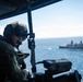 A military aircrew member wearing a flight helmet and tactical gear is positioned at an open door or window of an aircraft, appearing to operate or observe through a weapon or optical device. In the background, a naval vessel — consistent with a U.S. Navy amphibious transport dock ship — is visible on the open ocean. The image appears to document an aerial maritime patrol or training exercise.