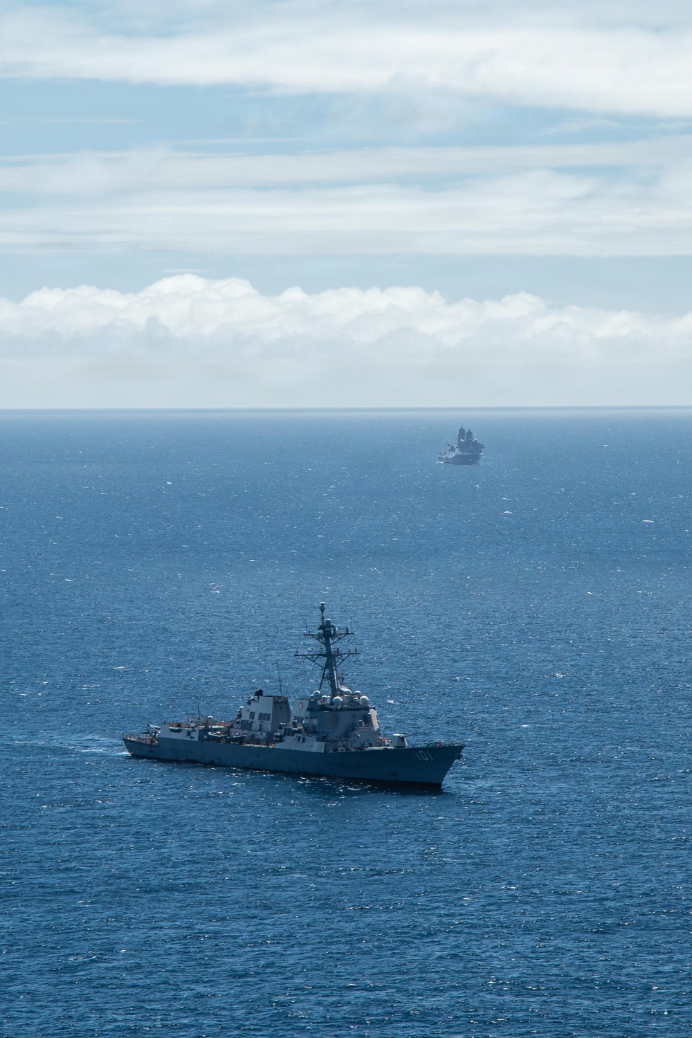 An aerial or elevated photograph showing two naval vessels underway on open ocean. The foreground vessel is a US Navy Arleigh Burke-class guided-missile destroyer bearing the hull number '101' on its bow. A second, larger vessel is visible in the distant background near the horizon.