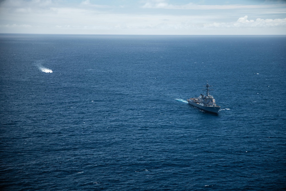 An aerial photograph shows a large gray naval warship underway in open ocean, consistent with a US Navy guided-missile cruiser or destroyer based on its silhouette and superstructure. The vessel is moving through deep blue water, leaving a visible white wake. In the upper left of the frame, a smaller vessel or object is also visible creating a wake, suggesting possible coordinated naval activity.