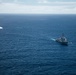 An aerial photograph shows a large gray naval warship underway in open ocean, consistent with a US Navy guided-missile cruiser or destroyer based on its silhouette and superstructure. The vessel is moving through deep blue water, leaving a visible white wake. In the upper left of the frame, a smaller vessel or object is also visible creating a wake, suggesting possible coordinated naval activity.