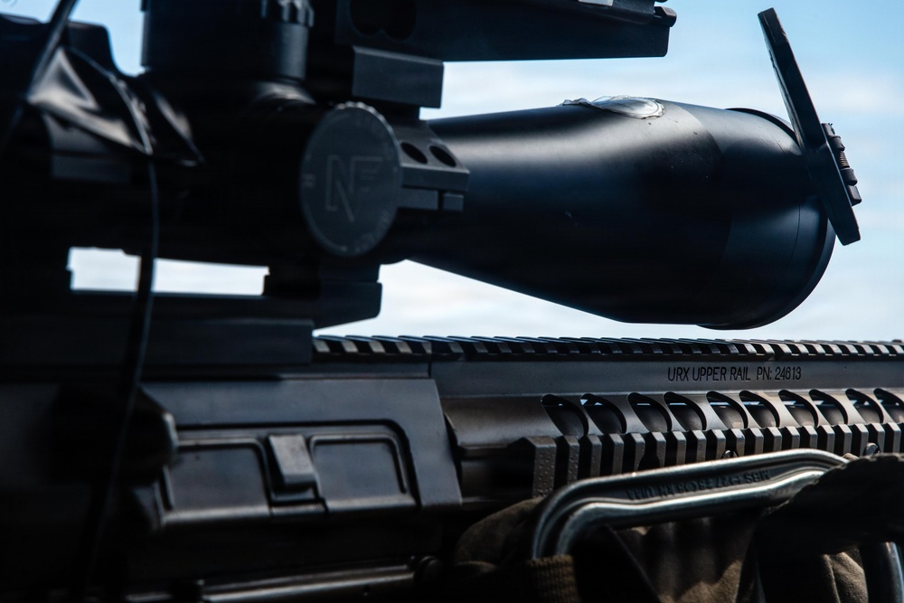 Close-up photograph of a tactical rifle platform with a large optical scope mounted on a Picatinny rail system. The image shows detailed markings on the upper rail and various accessories attached to the firearm. The shot is taken from a low angle against a partly cloudy sky background.