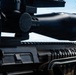 Close-up photograph of a tactical rifle platform with a large optical scope mounted on a Picatinny rail system. The image shows detailed markings on the upper rail and various accessories attached to the firearm. The shot is taken from a low angle against a partly cloudy sky background.