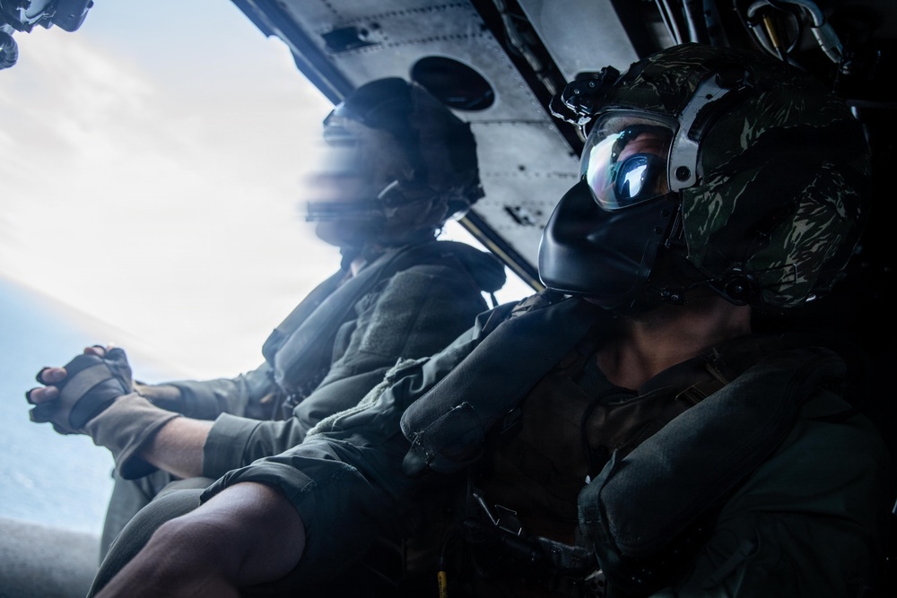 Two military personnel are seated inside an open-door helicopter or aircraft, likely during a flight over water. The individual in the foreground is wearing a flight helmet with a reflective visor and oxygen/communication mask, along with camouflage gear. The image captures an in-flight operational or training scenario with the open door revealing sky and ocean below.