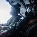 Two military personnel are seated inside an open-door helicopter or aircraft, likely during a flight over water. The individual in the foreground is wearing a flight helmet with a reflective visor and oxygen/communication mask, along with camouflage gear. The image captures an in-flight operational or training scenario with the open door revealing sky and ocean below.