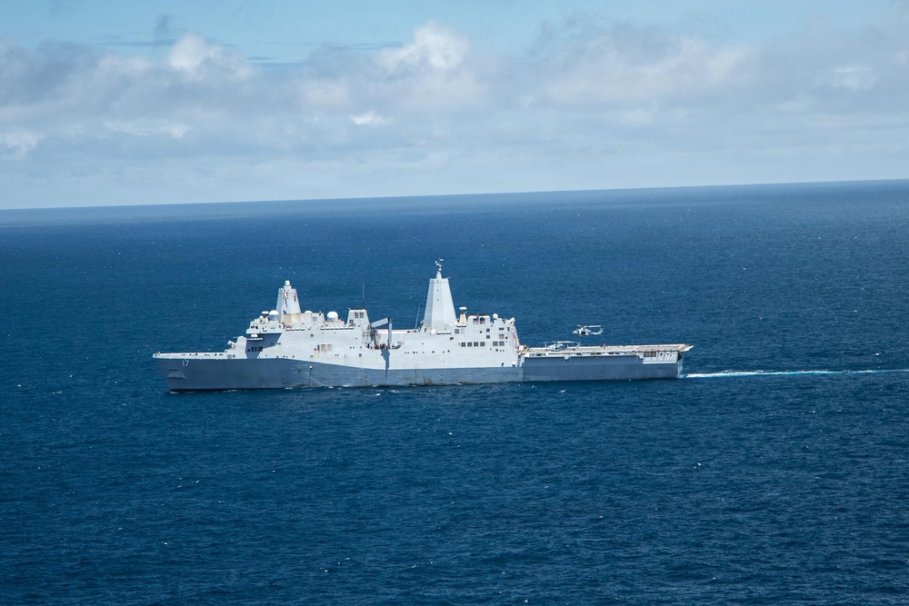 An aerial photograph of a large gray naval warship underway in open ocean. The vessel bears the hull number '17' on its bow and features a flat flight deck at the stern with what appears to be a helicopter either landing or departing. The ship is leaving a visible white wake as it travels through deep blue waters under a partly cloudy sky.