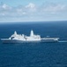 An aerial photograph of a large gray naval warship underway in open ocean. The vessel bears the hull number '17' on its bow and features a flat flight deck at the stern with what appears to be a helicopter either landing or departing. The ship is leaving a visible white wake as it travels through deep blue waters under a partly cloudy sky.