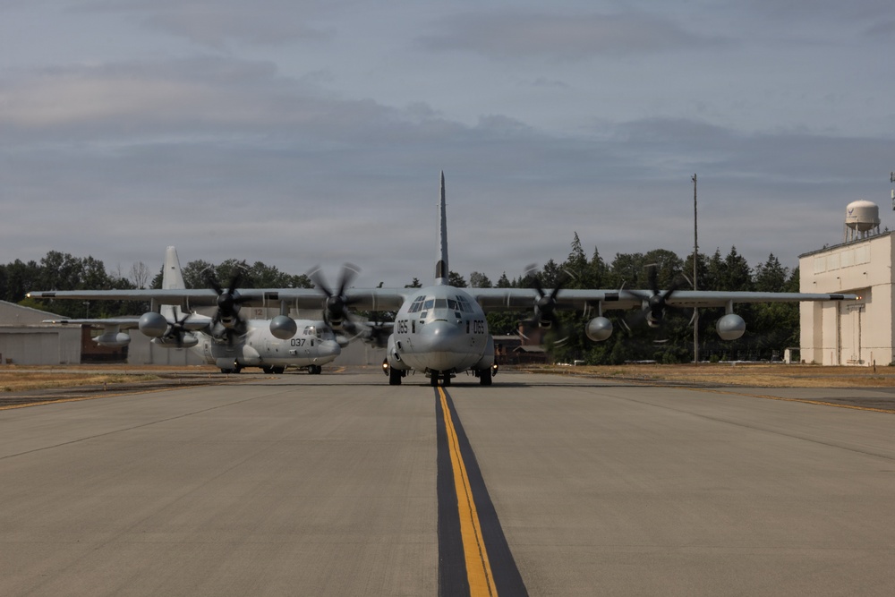 Two military turboprop aircraft, appearing to be C-130 Hercules variants, are taxiing on a military airfield tarmac. The foreground aircraft bears the number '065' and the background aircraft shows '037', both in gray military livery with spinning propellers indicating active engines. The setting appears to be a U.S. military installation with hangars, a water tower, and tree line visible in the background.