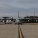 Two military turboprop aircraft, appearing to be C-130 Hercules variants, are taxiing on a military airfield tarmac. The foreground aircraft bears the number '065' and the background aircraft shows '037', both in gray military livery with spinning propellers indicating active engines. The setting appears to be a U.S. military installation with hangars, a water tower, and tree line visible in the background.