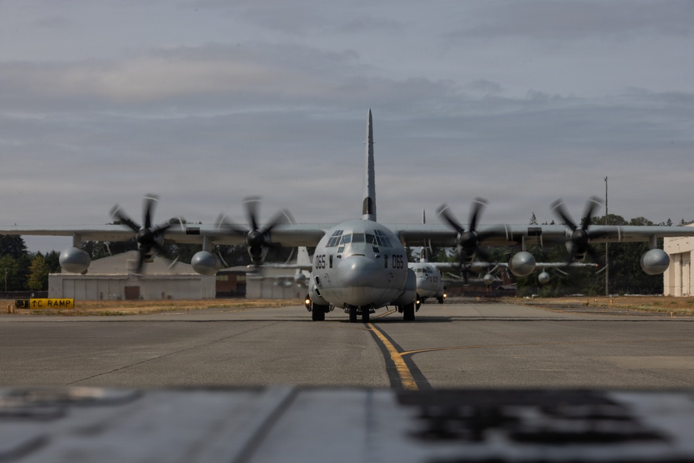 A military turboprop transport aircraft, identified as a C-130 Hercules with tail number 065, is taxiing on a runway or taxiway at a military airfield. All four propellers are spinning, visible as motion-blurred discs, indicating the engines are running at operational speed. The aircraft is photographed head-on from a low angle, with airfield infrastructure and trees visible in the background.