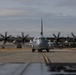 A military turboprop transport aircraft, identified as a C-130 Hercules with tail number 065, is taxiing on a runway or taxiway at a military airfield. All four propellers are spinning, visible as motion-blurred discs, indicating the engines are running at operational speed. The aircraft is photographed head-on from a low angle, with airfield infrastructure and trees visible in the background.